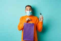 冠状病毒流感大流行生活方式概念男人。脸面具显示购物袋手洗手液站绿松石背景