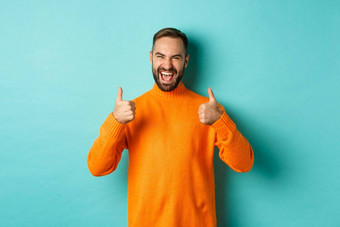 超级好满意快乐男人。显示拇指同意赞美优秀的工作很高兴站橙色毛衣光蓝色的背景