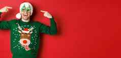 图像兴奋微笑的家伙指出聚会，派对眼镜庆祝一年站快乐的绿色毛衣圣诞老人他红色的背景