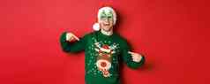 图像快乐男人。聚会，派对眼镜圣诞老人他指出圣诞节毛衣微笑站红色的背景