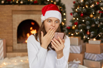 惊讶女人阅读电话内容圣诞节他看到令人惊讶的事物涵盖了口棕榈设备的屏幕大眼睛摆姿势生活房间壁炉x-mas树