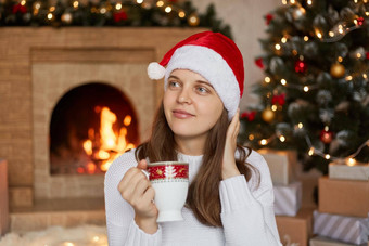 年轻的沉思的女人圣诞老人他圣诞节树壁炉首页<strong>如</strong>痴<strong>如</strong>梦持有杯咖啡<strong>茶</strong>穿白色温暖的毛衣