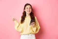美丽的现代女孩持有智能手机指出手指显示促销文本Copyspace移动电话站粉红色的背景