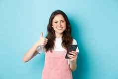 肖像美丽的快乐女孩显示拇指持有智能手机在线购物使订单互联网满意赞扬好应用程序蓝色的背景