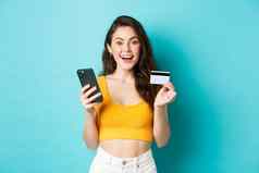 年轻的快乐女人使在线订单购物信贷卡智能手机惊讶相机站蓝色的背景