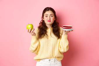 年轻的可爱的女人犹豫美味的一块<strong>蛋糕甜点</strong>绿色健康的苹果饮食站优柔寡断粉红色的背景