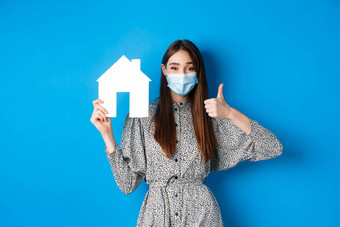 真正的房地产科维德流感大流行概念快乐的女人医疗面具显示纸房子断路拇指推荐机构房地产经纪人网站蓝色的背景