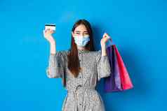 科维德流感大流行生活方式概念漂亮的女模型医疗面具欢乐购物折扣持有塑料信贷卡袋站快乐蓝色的背景