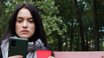 美丽的年轻的高加索人女人外套纸杯外卖咖啡采取自拍采取图片博客智能手机在户外公园坐着板凳上