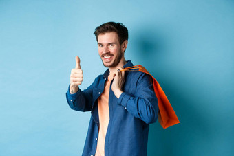 英俊的年轻的的家伙购物袋显示拇指微笑赞扬好促销活动提供站蓝色的背景