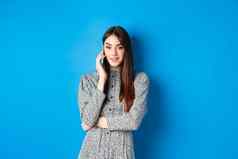 时尚的年轻的女人会说话的电话使外卖订单智能手机站衣服蓝色的背景