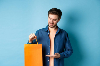 年轻的微笑的家伙显示橙色购物袋展示礼物站蓝色的背景