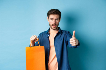 很酷的的家伙显示拇指持有橙色购物袋站蓝色的背景