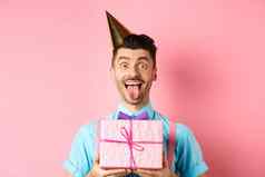假期庆祝活动概念有趣的的家伙庆祝生日穿聚会，派对他持有生日礼物显示舌头快乐脸粉红色的背景
