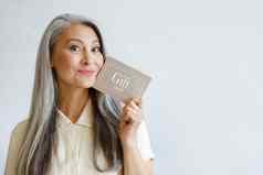 快乐的灰色头发的亚洲女人持有礼物卡摆姿势光灰色背景