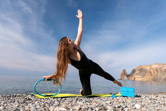 年轻的女人长头发健身教练黑色的运动服装紧身裤上衣伸展运动瑜伽席魔法普拉提环海阳光明媚的一天女健身瑜伽例程概念
