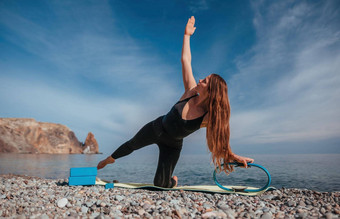年轻的女人长头发健身教练黑色的运动服装紧身裤上衣伸展运动瑜伽席魔法普拉提环海阳光明媚的一天女健身瑜伽例程概念