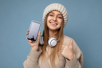 有吸引力的积极的微笑年轻的女人穿时尚的休闲装孤立的色彩鲜艳的背景墙持有显示移动电话空屏幕断路穿白色蓝牙耳机有趣的相机显示拇指手势