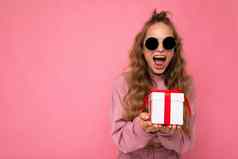 漂亮的惊讶震惊了快乐年轻的金发女郎卷曲的女人孤立的粉红色的背景墙穿粉红色的体育运动衣服太阳镜持有礼物盒子相机