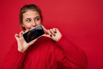照片美丽的可爱的年轻的女人穿温暖的<strong>红</strong>色的毛衣孤立的<strong>红</strong>色的<strong>背景墙</strong>持有智能手机显示电话空显示模型一边
