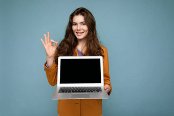 <strong>照片</strong>美丽的微笑满意快乐年轻的女人持有电脑移动PC相机穿黄色的夹克显示手势孤立的蓝色的<strong>墙背景</strong>