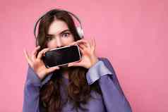 照片漂亮的积极的年轻的浅黑肤色的女人卷曲的女人穿紫色的作物前孤立的粉红色的背景墙持有建议移动电话空显示蓝牙耳机相机