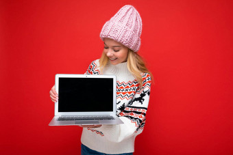 照片拍摄美丽的迷人的微笑金发女郎年轻的女人持有电脑移动PC空监控屏幕模拟复制空间穿针织冬天他毛衣上网本屏幕孤立的红色的墙背景