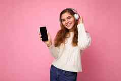 有吸引力的积极的微笑年轻的女人穿时尚的休闲装孤立的色彩鲜艳的背景墙持有显示移动电话空屏幕断路穿白色蓝牙耳机有趣的相机
