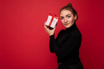 迷人的积极的年轻的浅黑肤色的女人女人孤立的红色的背景墙穿黑色的毛衣持有白色礼物盒子红色的丝带相机