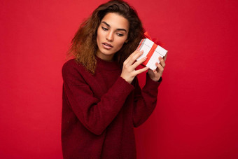 有吸引力的积极的集中年轻的褐色卷曲的女人孤立的红色的背景墙穿红色的毛衣持有礼物盒子猜一猜惊喜