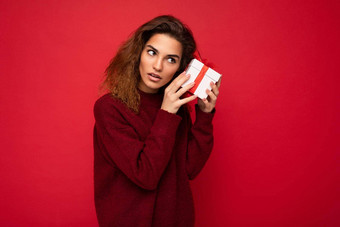 照片拍摄有吸引力的深思熟虑的集中年轻的浅黑肤色的女人卷曲的女人孤立的红色的背景墙穿红色的毛衣持有礼物盒子一边思考