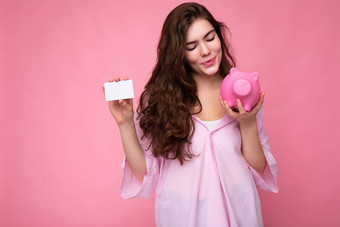 有吸引力的快乐年轻的浅黑肤色的女人女人穿衬衫孤立的粉红色的背景空空间持有粉红色的猪钱盒子信贷卡模型
