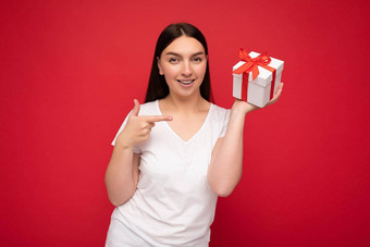 照片美丽的积极的微笑年轻的浅黑肤色的女人女人孤立的红色的背景墙穿白色休闲t恤模型持有白色礼物盒子红色的丝带相机指出手指现在