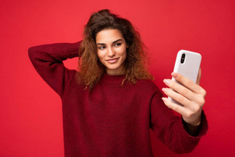 美丽的年轻的女人卷曲的头发穿黑暗红色的毛衣孤立的红色的背景墙持有聪明的电话电话屏幕采取自拍