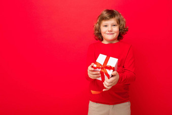 照片拍摄快乐的微笑金发女郎卷曲的男孩孤立的红色的背景墙穿红色的毛衣持有礼物盒子相机