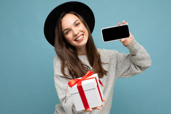 拍摄漂亮的微笑积极的年轻的褐色女人孤立的<strong>蓝色</strong>的<strong>背景墙</strong>穿时尚的黑色的他灰色毛衣持有礼物盒子显示智能手机屏幕显示模型相机