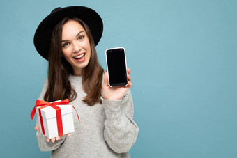 拍摄漂亮的微笑积极的年轻的浅黑肤色的女人女人孤立的蓝色的背景墙穿时尚的黑色的他灰色毛衣持有礼物盒子显示智能手机屏幕显示模型相机