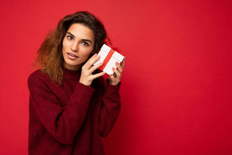 美丽的深思熟虑的年轻的浅黑肤色的女人卷曲的女人孤立的红色的背景墙穿红色的毛衣持有礼物盒子相机