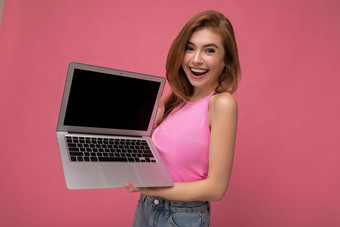 快乐微笑美丽的年轻的金发碧眼的女人持有电脑移动PC穿粉红色的作物前相机孤立的粉红色的背景