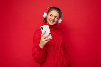 照片美丽的快乐年轻的浅黑肤色的女人女人穿红色的毛衣孤立的红色的背景墙持有移动电话穿蓝牙耳机有趣的