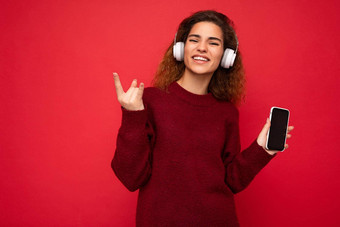 拍摄有吸引力的积极的微笑年轻的褐色卷曲的女人穿黑暗红色的毛衣孤立的红色的背景墙穿白色无线耳机听音乐显示移动电话空屏幕断路相机显示岩石卷标志