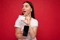 照片有吸引力的快乐年轻的浅黑肤色的女人女人穿休闲白色t恤孤立的红色的背景空空间持有手移动电话显示智能手机空屏幕模型信贷卡一边