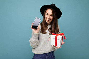 拍摄漂亮的微笑积极的年轻的浅黑肤色的女人女人孤立的蓝色的背景墙穿时尚的黑色的他灰色毛衣持有礼物盒子显示智能手机屏幕显示模型相机
