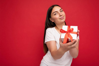 照片美丽的积极的年轻的浅黑肤色的女人女人孤立的红色的背景墙穿白色休闲t恤模型持有白色礼物盒子红色的丝带相机