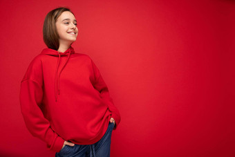 照片拍摄有吸引力的快乐积极的微笑浅黑肤色的女人女少年穿时尚的红色的连帽衫站孤立的红色的背景墙一边