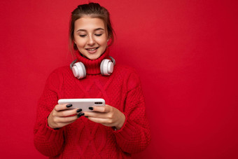 有吸引力的快乐年轻的褐色女人穿红色的毛衣孤立的红色的背景墙持有移动电话冲浪互联网穿蓝牙耳机智能手机