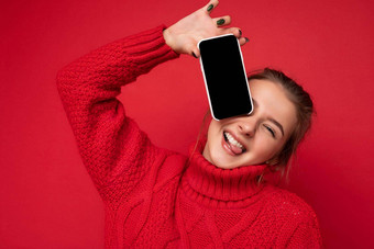 照片美丽的可爱的微笑年轻的女人穿温暖的红色的毛衣孤立的红色的背景墙持有智能手机显示电话空显示模型相机
