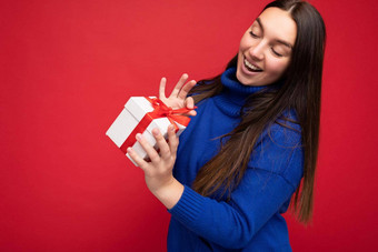 拍摄有吸引力的快乐微笑年轻的浅黑肤色的女人女人孤立的红色的背景墙穿蓝色的休闲毛衣持有白色礼物盒子红色的丝带相机