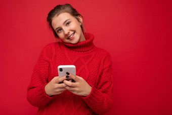 有吸引力的可爱的微笑年轻的女人穿温暖的红色的毛衣孤立的红色的背景墙持有智能手机沟通在线互联网相机