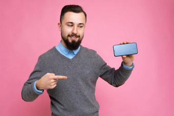 英俊的快乐很酷的年轻的浅黑肤色的女人不剃须的男人。beardwearing时尚的灰色毛衣蓝色的衬衫站孤立的粉红色的<strong>背景墙</strong>持有智能手机显示电话空屏幕显示设备